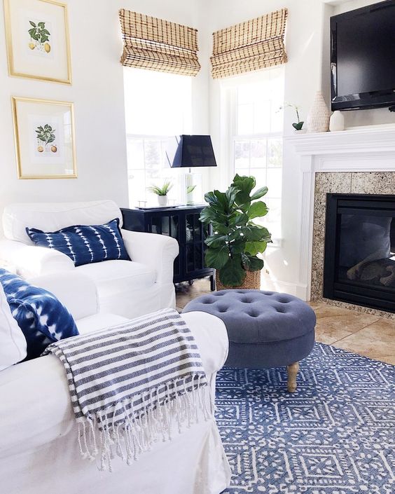 a cozy beach living space with white furniture, a blue ottoman, a printed rug and woven Roman shades
