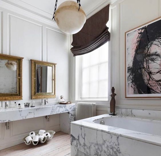 a gorgeous bathroom with a white marble vanity and tub, a statement artwork, mirrors in vintage frames and shades