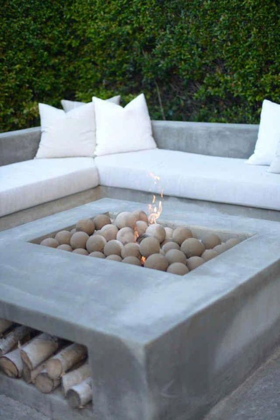 a minimalist outdoor fire pit with a built in concrete corner bench, with neutral upholstery and a matching concrete fire bowl with stones and firewood storage