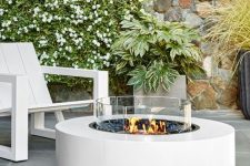 a modern outdoor fire pit with a stylish ethanol fire bowl in white and white wooden chairs plus greenery and blooms around