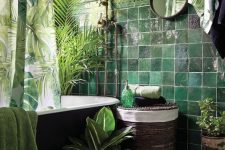 a moody tropical bathroom with green walls and dark green glossy tiles, a black tub, a woven stool, potted plants and tropical print textiles