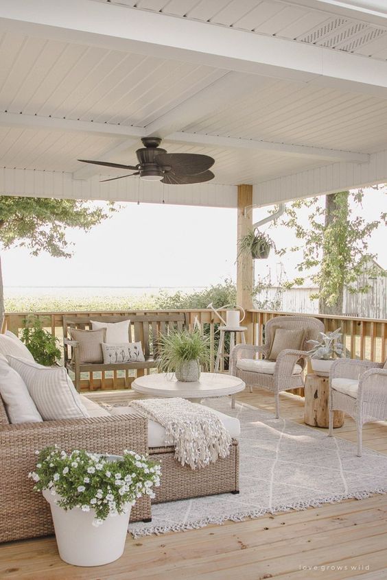 a neutral rustic seaside terrace with light colored wicker furniture, white upholstery and textiles and potted blooms and greenery