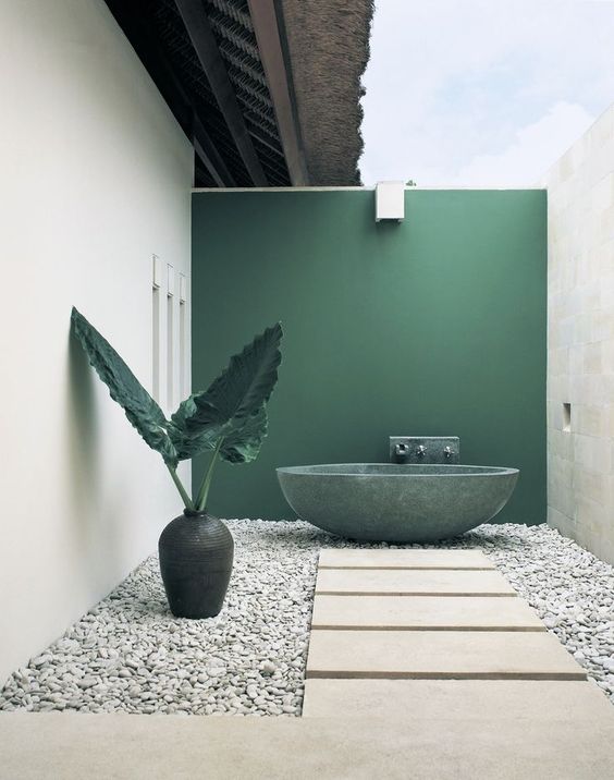 a refined minimalist outdoor bathroom with a green statement wall and bathtub, pebbles, stone tiles and a large vase