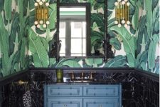a refined vintage tropical bathroom with banana leaf wallpaper, black marble tiles, a navy vanity and touches of gold