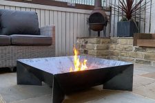 a stylish modern fire pit with wicker furniture, a black square metal fire pit, potted plants and a stone tile floor