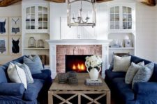 a traditional coastal living room in navy and white, with stripes, a brick clad fireplace, wooden beams and built-in shelves