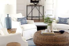 a welcoming coastal living room with a rattan ottoman, a striped rug, white furniture and blue lamps and pillows