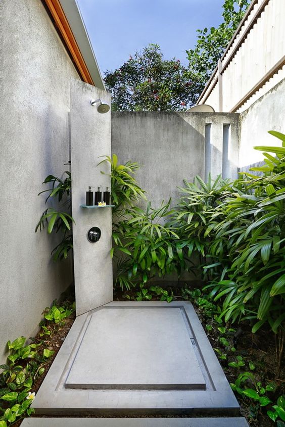 a welcoming outdoor shower with much greenery and a concrete shower zone with a stand