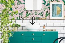 an airy and fun tropical bathroom with catchy wallpaper, a green vanity, cascading greenery and touches of gold