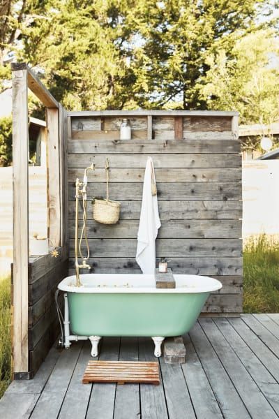 an outdoor rustic bathroom nook with weathered wood screens, a green tub, a pallet rug and some accessories