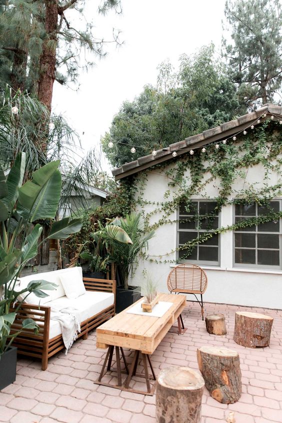 a boho meets rustic patio with wooden and wicker furniture, white textiles, potted greenery and tree stumps