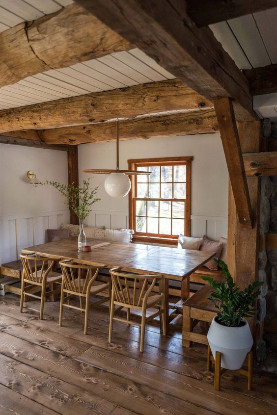 a cozy rustic dining space with stained wooden beams, a built in bench, a stained table and chairs, a lovely pendant lamp and greenery