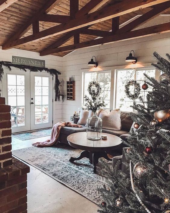 a cozy rustic living room with a round wooden table, a brick fireplace, a wooden ceiling with beams and an evergreen garland