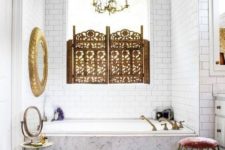 a neutral bathroom with a Moroccan feel, a gilded mirror, a boho rug, a beautiful screen on the window and a geometric stool