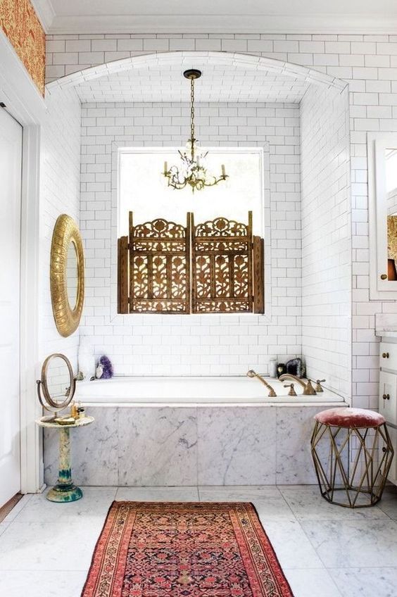 a neutral bathroom with a Moroccan feel, a gilded mirror, a boho rug, a beautiful screen on the window and a geometric stool