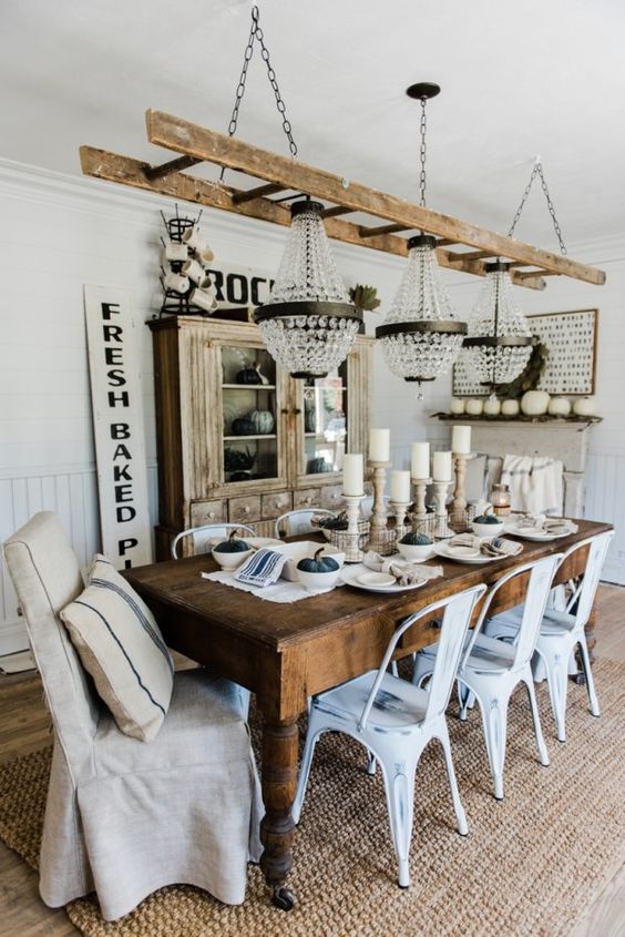 a neutral farmhouse dining room with a shabby chic buffet, a dark stained table, white metal chairs, a ladder with crystal chandeliers