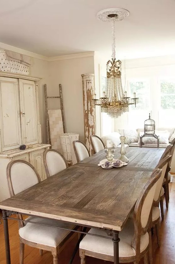 a neutral vintage dining room with a white storage unit, a stained dining table, neutral upholstered chairs, a crystal chandelier and some shabby chic decor