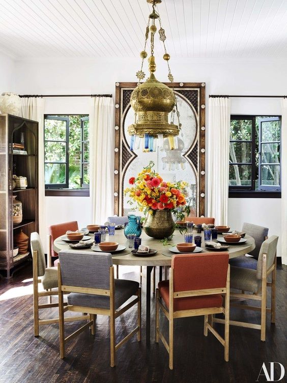 a pretty Moroccan-inspired dining room with a round table, muted color chairs, a bold metal pendant lamp and a large mirror in a carved wooden frame