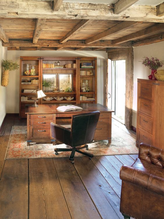 a rustic home office with a rough wooden ceiling, heavy wooden furniture, a vintage rug, a leather chair and some lamps