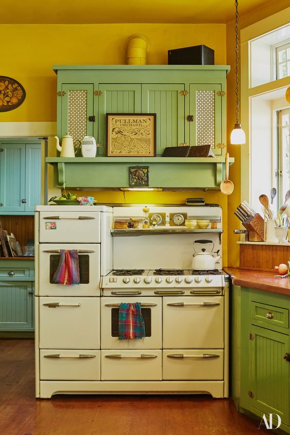 a vintage kitchen with mustard walls, vintage green cabinets, neutral appliances and pendant lamps