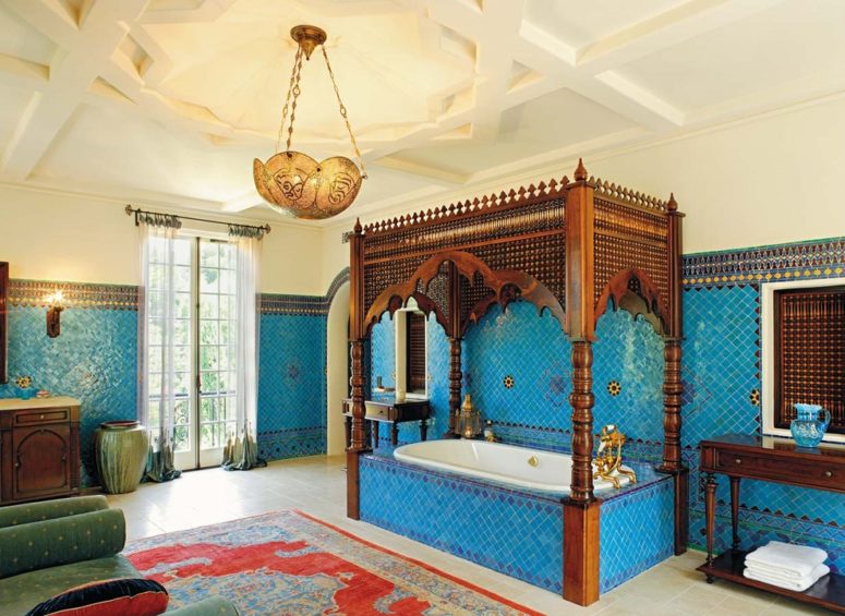 a bold blue Moroccan bathroom with a hanging lamp, a bathtub with a wooden frame on top and a bright rug and vases  (Chris Barrett Design)