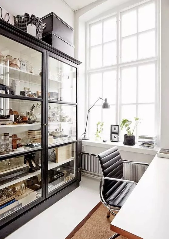 a catchy Scandinavian home office with a large glass storage unit, a white desk, a black chair, potted plants and artwork