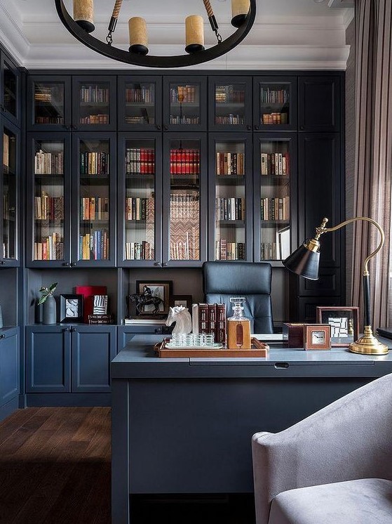 a chic blue home office, all the walls taken by storage units, a blue desk, a black chair and a catchy chandelier with candles