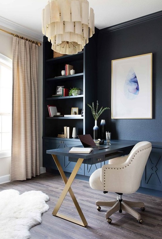 a chic home office with a blue wall taken by storage units, a statement artwork, a unique chandelier, a Murphy desk and a large white chair