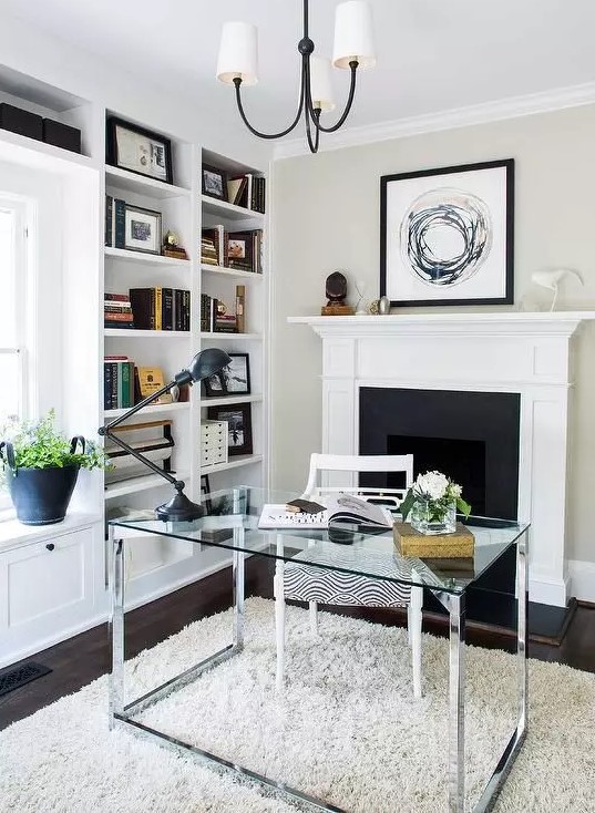 a chic modern neutral home office with built-in bookshelves, a fireplace, a glass desk, a neutral rug and a stylish chandelier