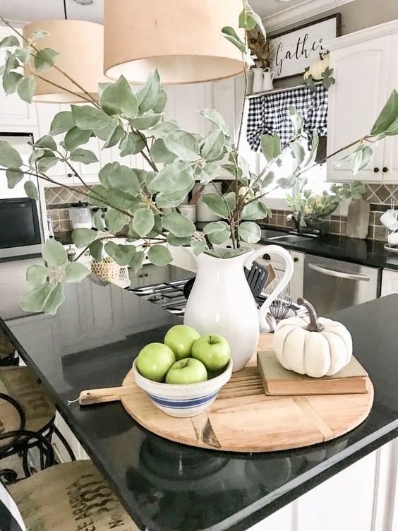 a cutting board with a book, a fake pumpkin, a bowl with green apples and a greenery arrangement in a white jug
