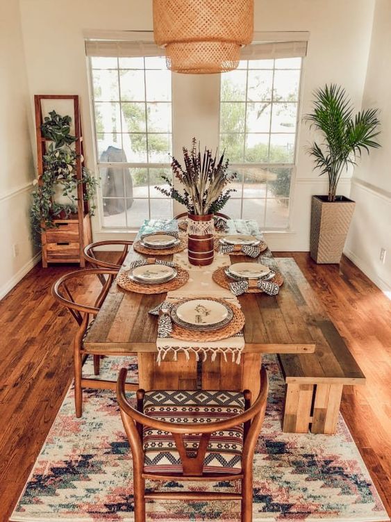 a gorgeous boho dining space with potted greenery, a boho rug, a wicker pendant lamp and warm-stained furniture