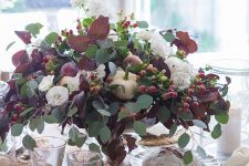a gorgeous fall centerpiece of eucalyptus, dark foliage, berries, white blooms and pumpkins and fruit