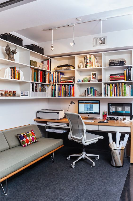 a mid-century modern home office with a large open storage corner unit with lights, a desk and a white chair, a grey sofa