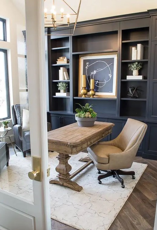 a modern farmhouse home office with a navy storage unit that takes a whole wall, a wooden desk, leather chairs and a refined gold chandelier