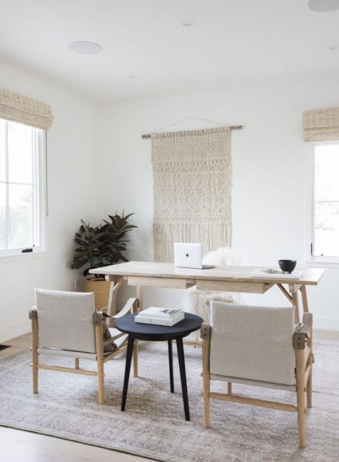 a neutral boho home office with a large macrame wall hanging, a simple wooden desk and neutral chairs plus wicker shades