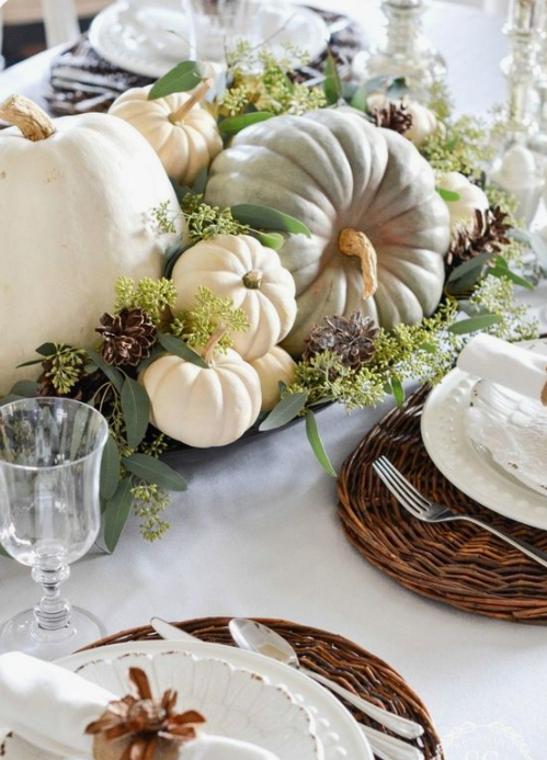a neutral fall table setting with a bowl with natural pumpkins, greenery and pinecones, woven placemats and white porcelain