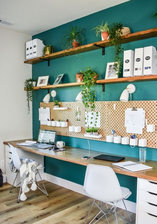 a shared home office with an emerald accent wall, open shelves, a pegboard, a shared desk with drawers and white Eames chairs