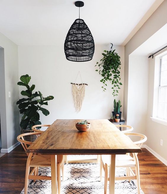 a simple boho dining room with a macrame hanging, a black wicker pendant lamp, wooden furniture and potted greenery and cacti