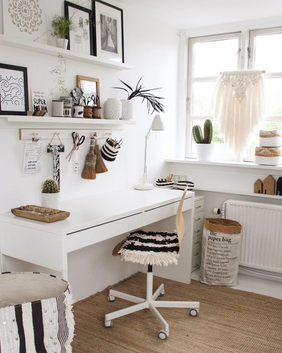 a white boho home office with open shelves, a white desk, a chair with a fringe cover, a boho ottoman, a macrame hanging and potted plants