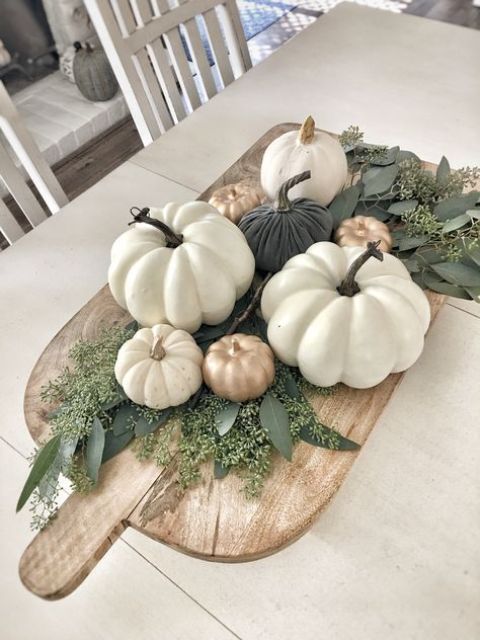 a wooden cutting board with greenery and fake pumpkins in white, gold and dark green as a fall centerpiece