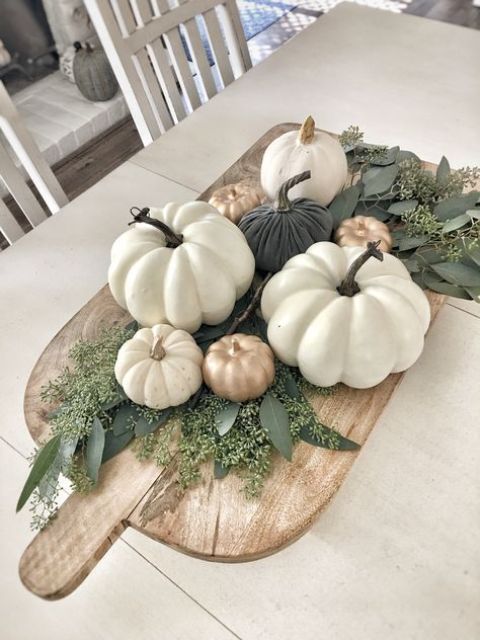 une planche à découper en bois avec de la verdure et de fausses citrouilles en blanc, or et vert foncé comme pièce maîtresse d'automne une planche à découper en bois avec de la verdure et de fausses citrouilles en blanc, or et vert foncé comme pièce maîtresse d'automne