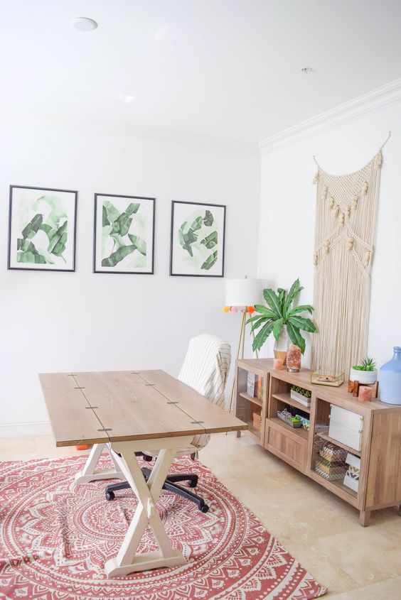an airy boho chic home office with a wooden desk and a storage unit, potted plants, a macrame hanging, a bright rug and a gallery wall