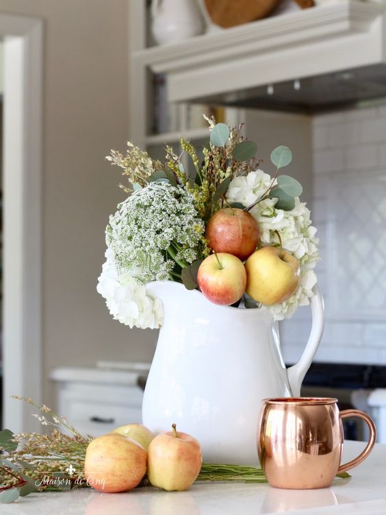 an arrangement of a white jug, white blooms, greenery, apples and a copper mug for simple and chic fall decor