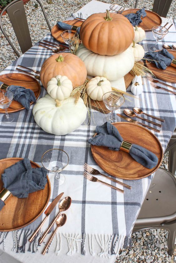 a cozy rustic fall tablescape with large pumpkins stacked, wheat, slate grey napkins and a plaid tablecloth, copper cutlery and wooden chargers