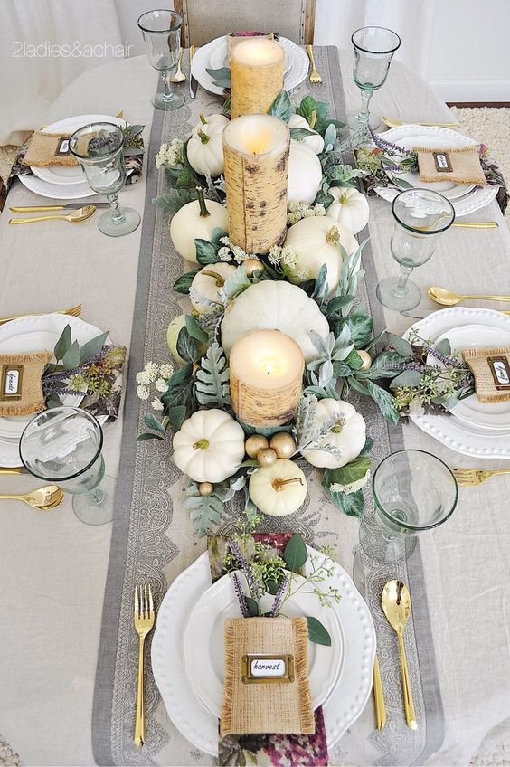 a fall tablescape with white pumpkins, candles wrapped with birch bark, gold cutlery and burlap pockets with blooms and greenery