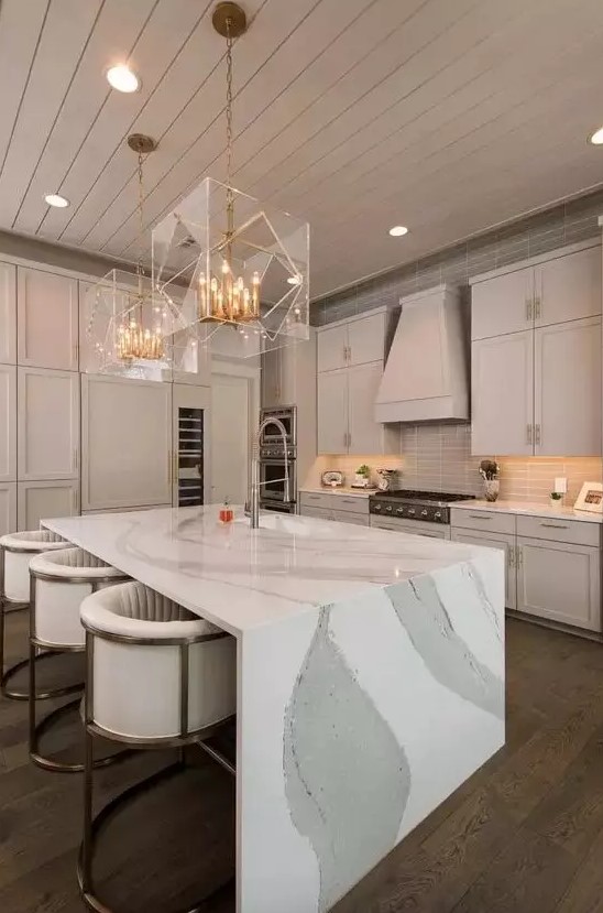 a refined neutral-colored kitchen with a jaw-dropping kitchen island with a waterfall countertop and stunning gold and sheer glass chandeliers