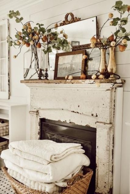 a shabby chic and rustic fall mantel decorated with mirrors, an artwork, branches with pears in vases and blankets in a basket