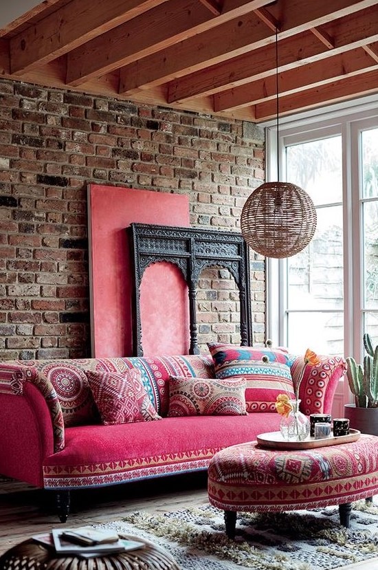 a boho living room with a brick wall, a pink sofa with printed pillows, a matching ottoman, a rattan pendant lamp and a Moroccan rug