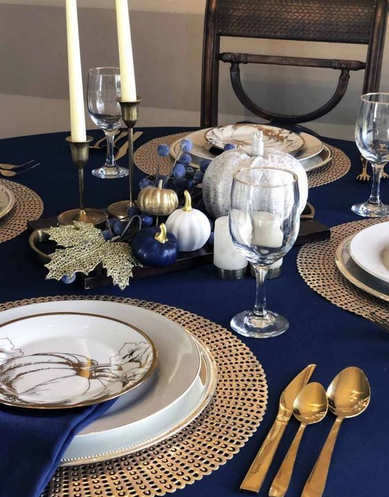 a bold Thanksgiving table with gold cutlery, woven chargers, printed plates, blue, gold and white pumpkins and candles