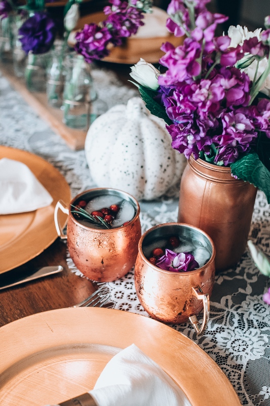 a bright Thanksgiving tablescape with a lace runner, bold purple blooms, spaeckled pumpkins, copper mugs and chargers